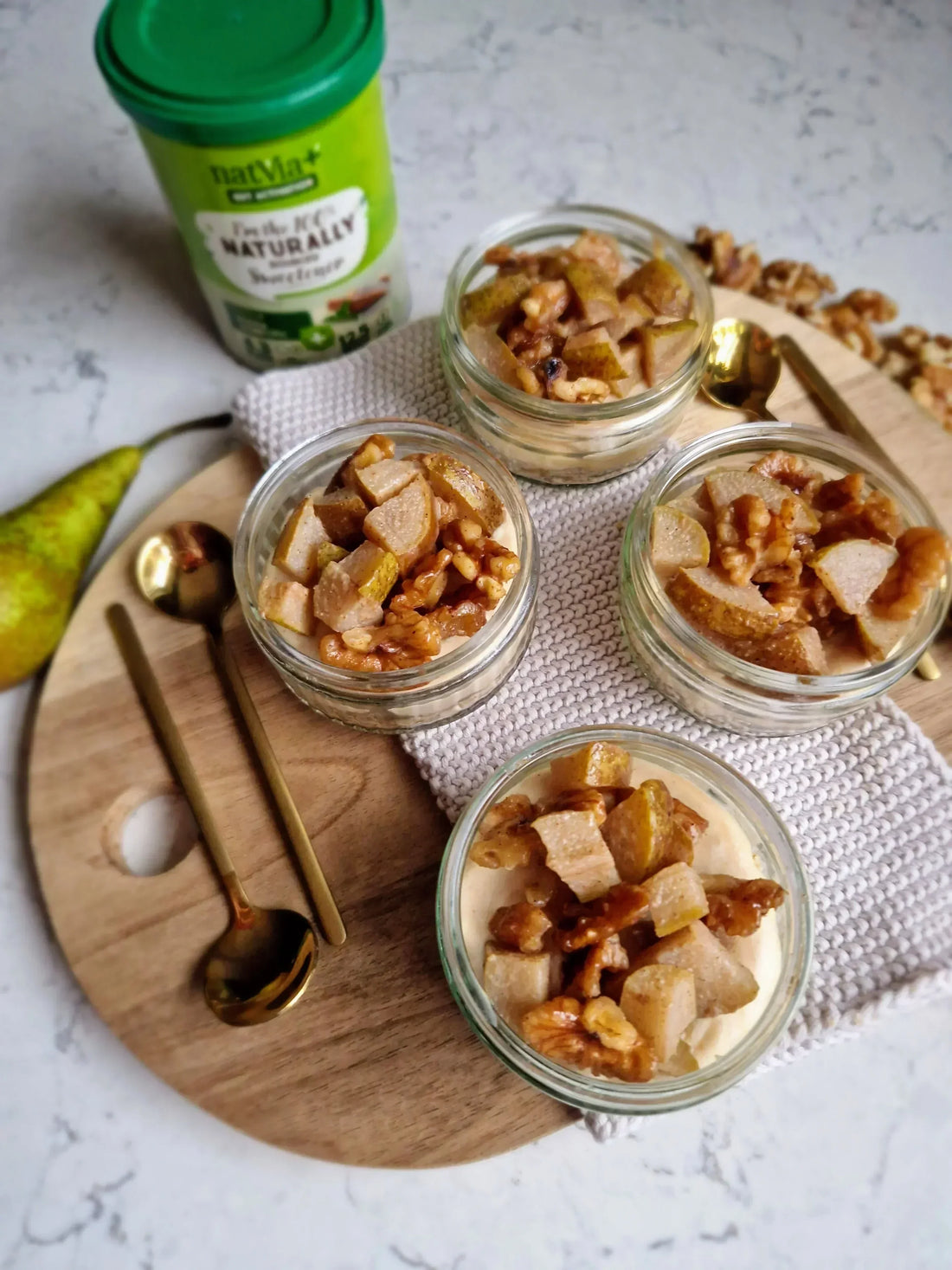 Four glass ramekins filled with overnight oat pots topped with sautéed diced pears and walnut pieces, arranged on a wooden serving board with gold spoons, a fresh pear, and a canister of Natvia Gut Activation Sweetener in the background.