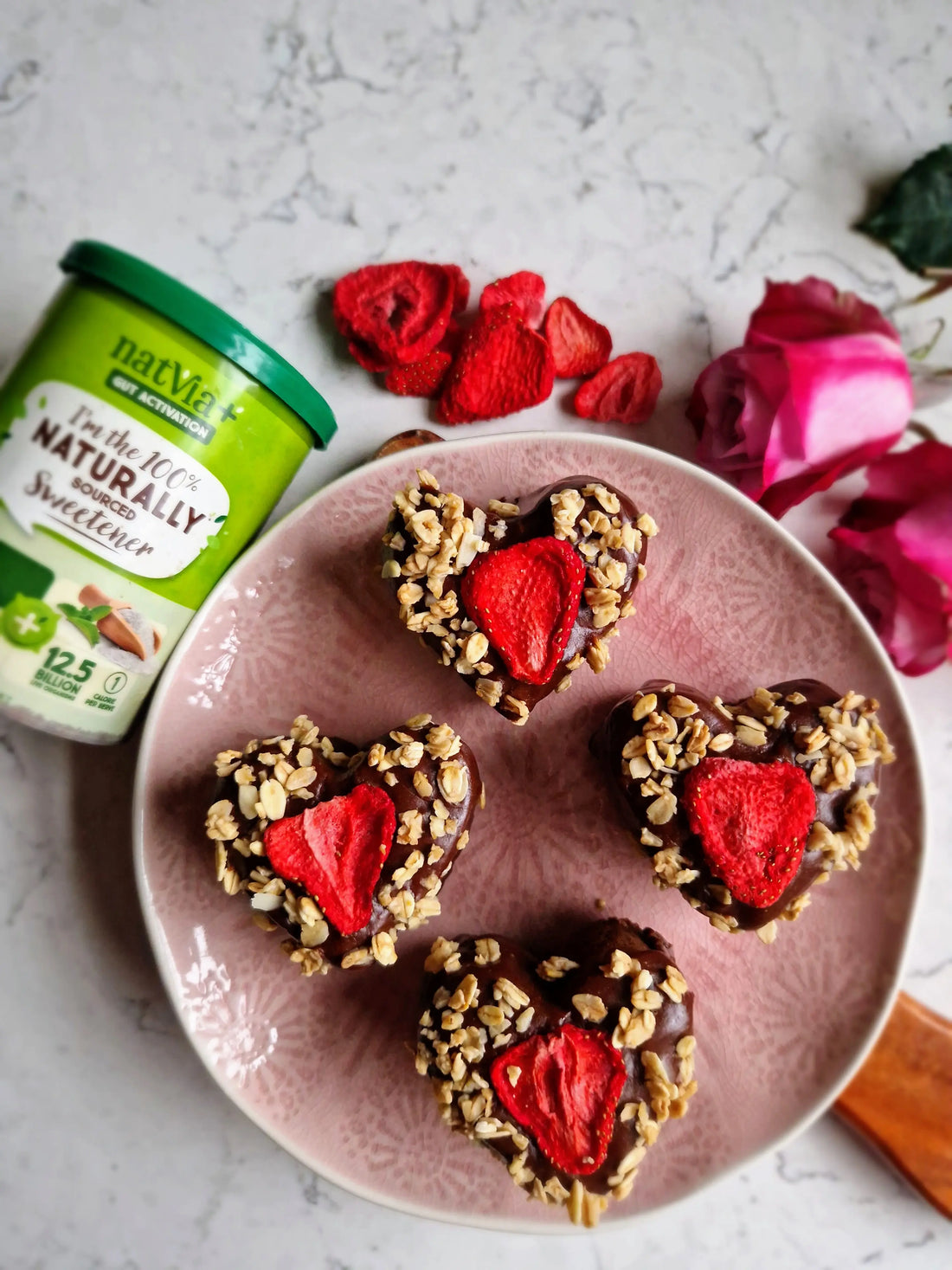 Four heart-shaped chocolate cakes on a wire rack, glazed with dark chocolate icing and topped with freeze-dried strawberries and granola sprinkles.