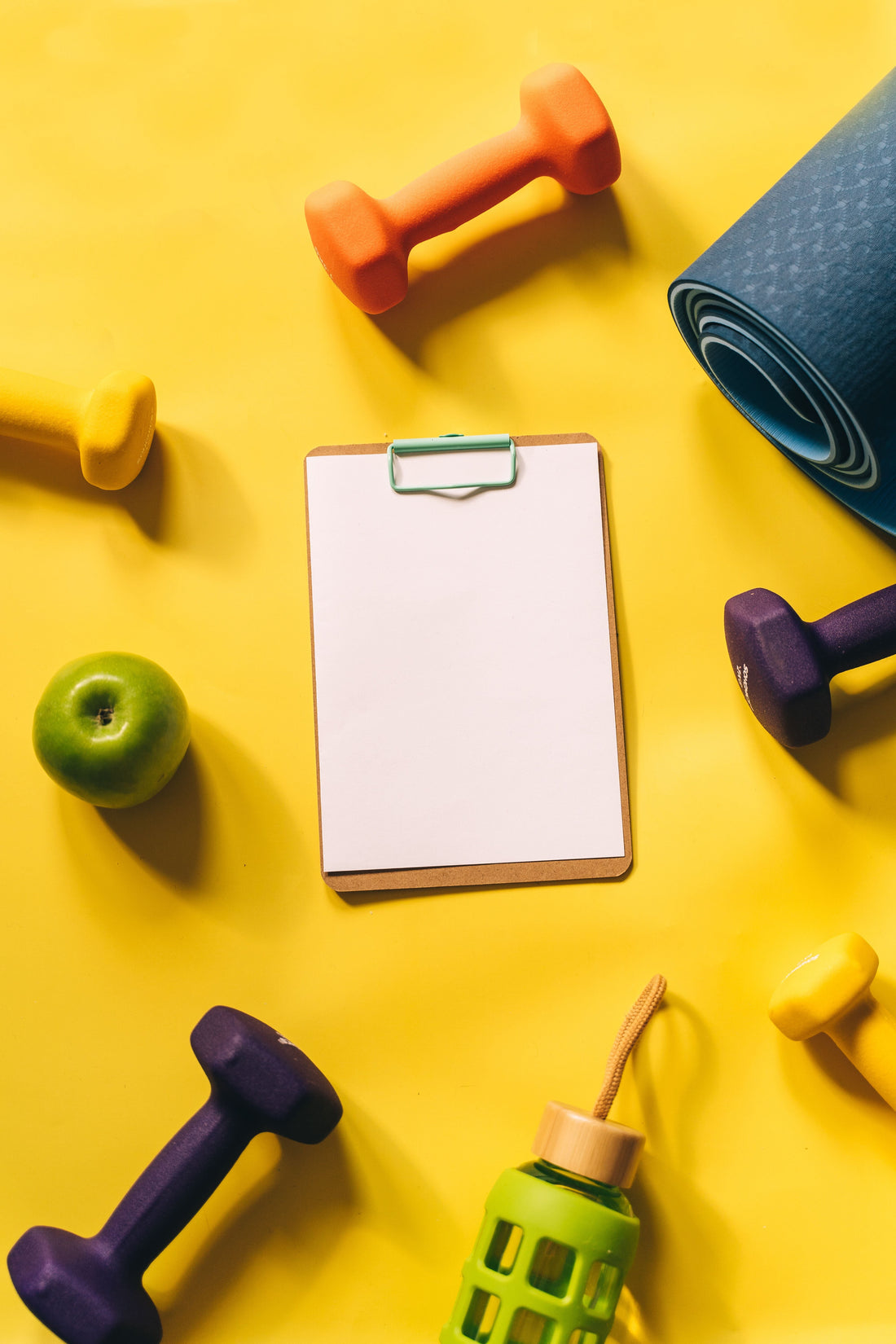 apple, colourful dumbbells, mat, and a notepad spread throughout a yellow background.
