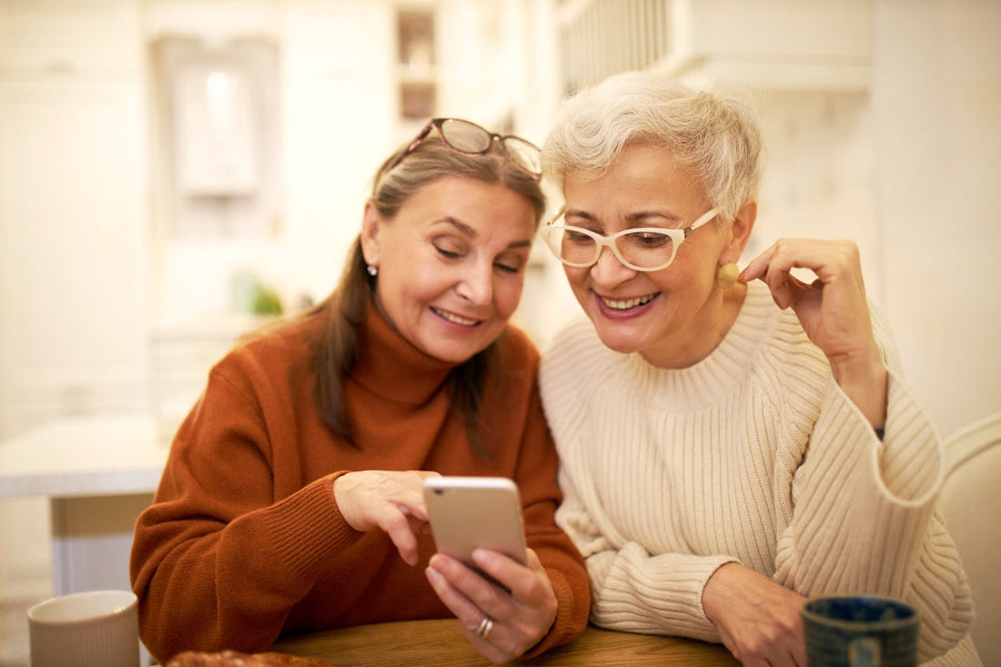 Two stylish retired female friends enjoying modern electronic gadget, sitting at cafe, watching pictures on mobile, recollecting good old days. Elderly women shopping online using smart phone.