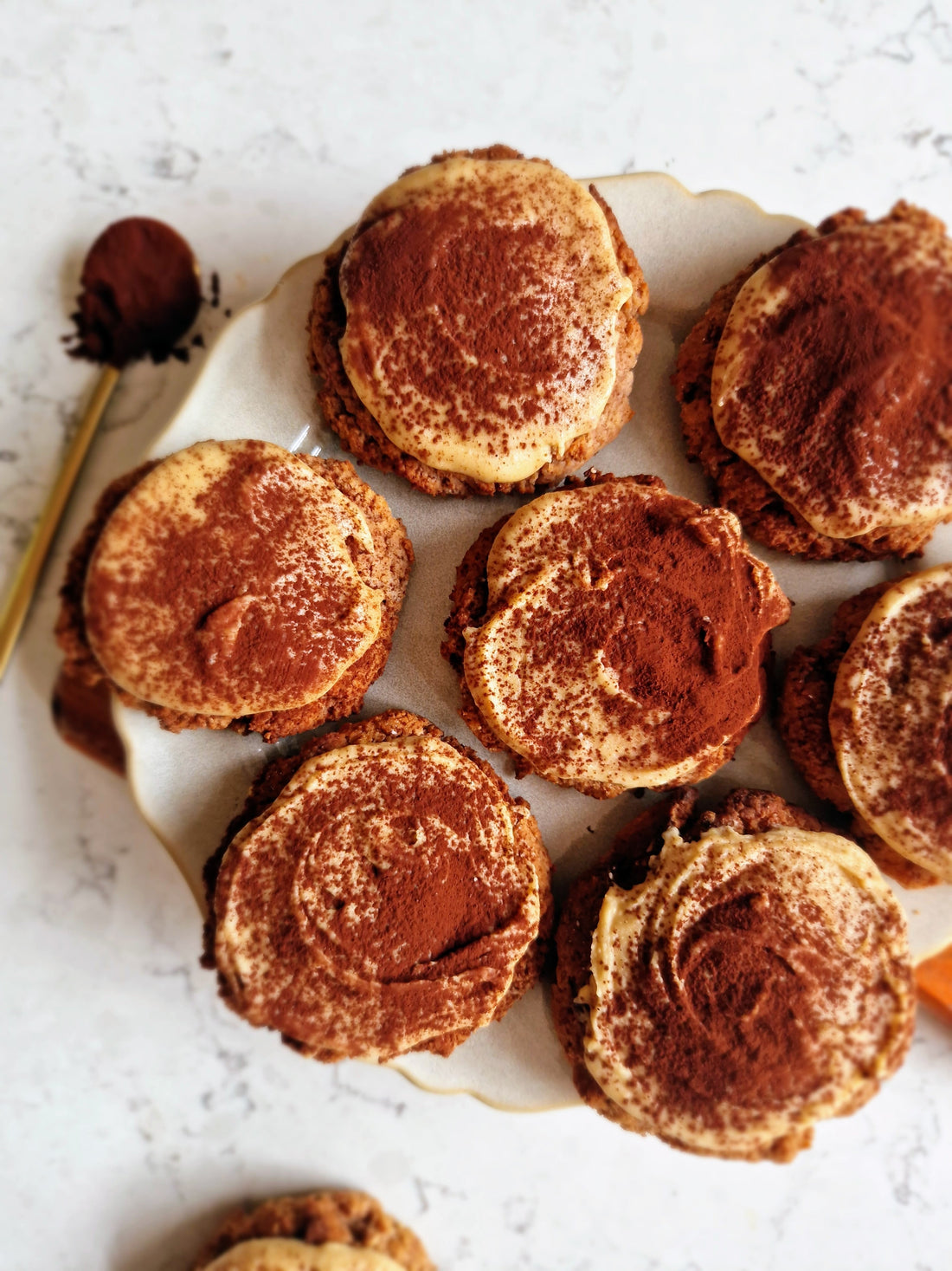 a plate full of tiramisu cookies topped with frosting and chocolate powder. And a spoon of chocolate powder on the side.