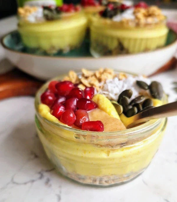No-bake turmeric pomegranate cheesecake pots in glass ramekins, topped with pomegranate seeds, granola, pumpkin seeds, and coconut, next to a spoon.