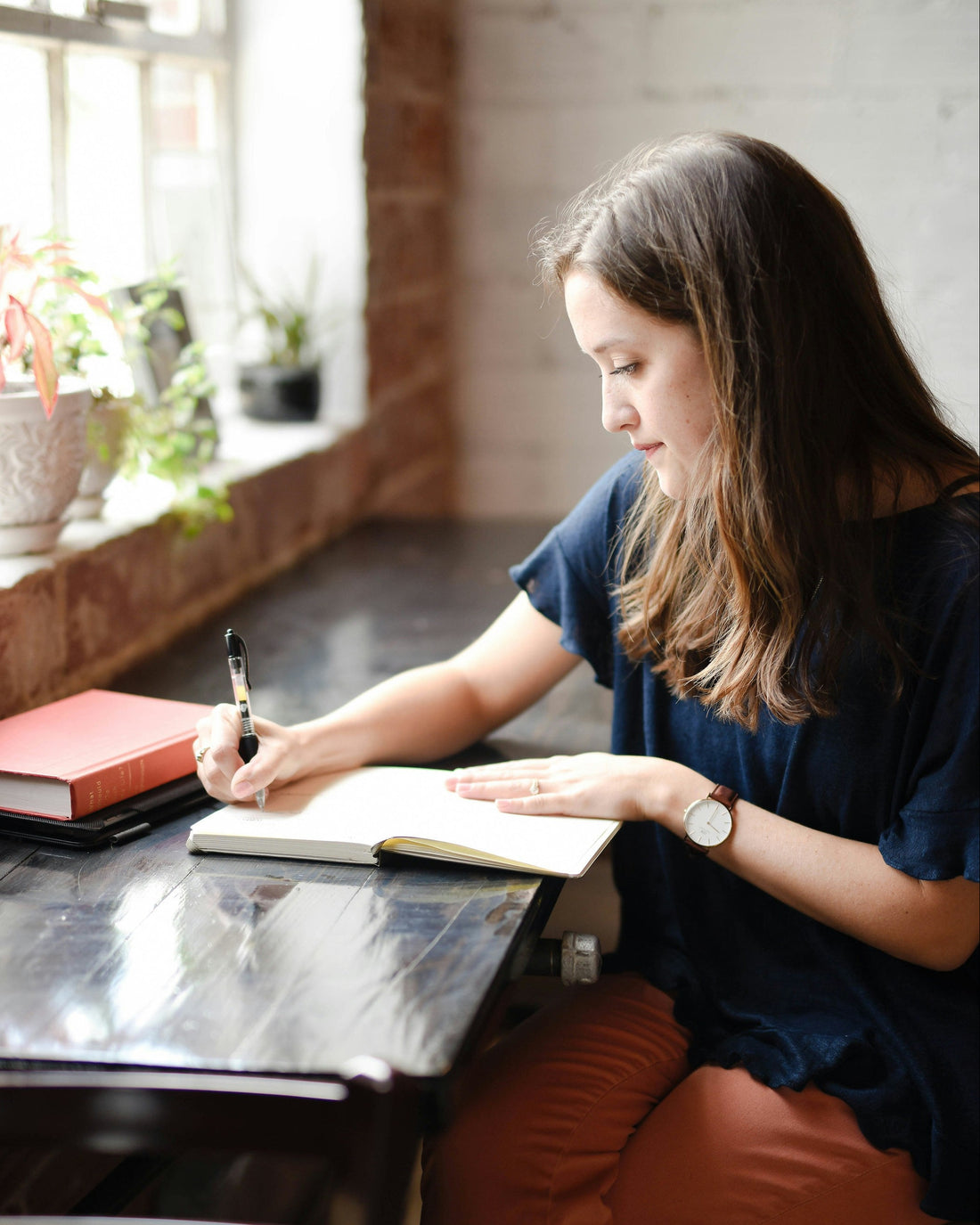 Woman sitting at a table writing in a notebook with a cup and books on the table.